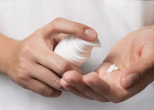 Close up of woman squeezing hand cream on her finger and holding a cosmetic bottle in other hand against grey. Beauty treatment or handcare procedure in a spa salon