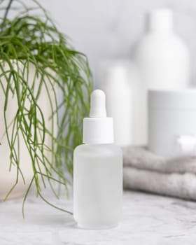 Glass dropper bottle on white marble against potted green plant and cosmetics in bathroom, close up, packaging mockup. Natural product for facecare - serum or essence