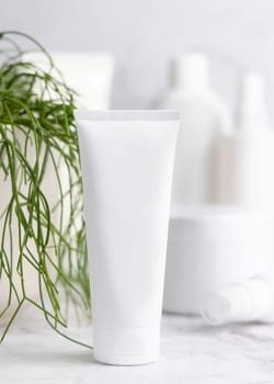 White cream squeeze tube on white marble table against green plant and cosmetic bottles in bathroom, close up, packaging mockup. Natural product for face care