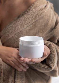 Close up of woman in light brown bathrobe holding opened cream jar in hands with natural manicure, mockup. Bodycare concept. White blank cosmetic container. Cropped Image