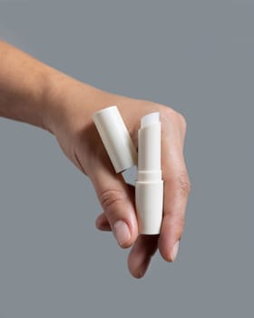 Female hand holding white opened lip balm stick with a lid against grey close up, cosmetic package mockup, copy space. Lip care beauty product with minimal composition