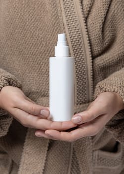 Close up of woman in light brown bathrobe with white cosmetic bottle, mockup. Hands with natural manicure holding blank spray bottle, beauty or spa procedures