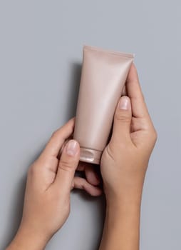 Female hands holding beige squeeze cream tube against grey close up, cosmetic package mockup, copy space. Skincare beauty product with minimal composition
