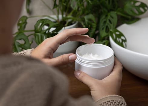 Close up of woman in brown bathrobe touching cream with finger and holding a cosmetic jar in other hand in bathroom against green plant and vessel basin. Shoulder view