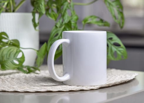 Closeup of 11 oz white blank coffee mug on grey table over blured green plant and modern interior, mockup. Template for your design or advertising with copy space