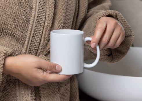 Close up of woman in light brown bathrobe 11 oz white blank coffee mug in hands, mockup. Hands with natural manicure. Template for your design with copy space