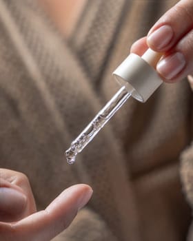 Close up of woman in brown bathrobe dropping essential oil on a finger and holding a pipette on other hand. Healthy hand skin and nails, natural manicure