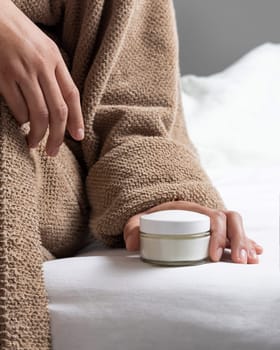 Woman in brown bathrobe puts white cream jar on white bedsheet closeup, mockup. Bodycare concept. Organic cosmetics for body and face care, beauty and spa procedures