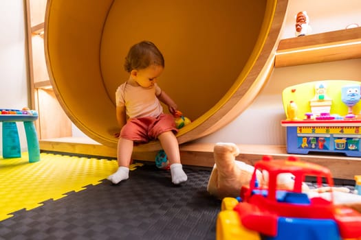 Toddler sitting in a round play structure surrounded by colorful toys and activity centers in a creative and fun indoor playroom