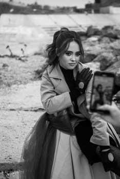 brunette girl in beige coat sits on chair among rocks. she looks at camera and smiles. she has curly dark hair. she holds bouquet of chrysanthemums in her hands, she has black gloves on her hands