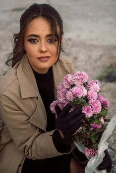 brunette girl in beige coat sits on chair among rocks. she looks at camera and smiles. she has curly dark hair. she holds bouquet of chrysanthemums in her hands, she has black gloves on her hands