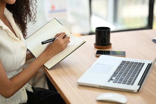 Cropped shot of female office worker writing in a notebook while working on laptop.