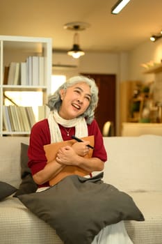 Calm senior woman in a red sweater sitting on couch and writing in journal.