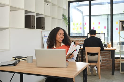 Confident African businesswoman presenting data on a clipboard while working on laptop.