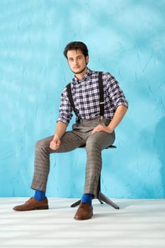 Handsome brunette in shirt and vest sitting on a chair against blue background portrait