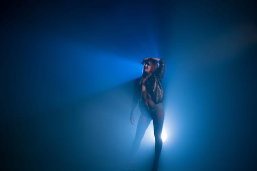 Brunette topless wearing black leather unbuttoned jacket and jeans shorts full-length view in blue light