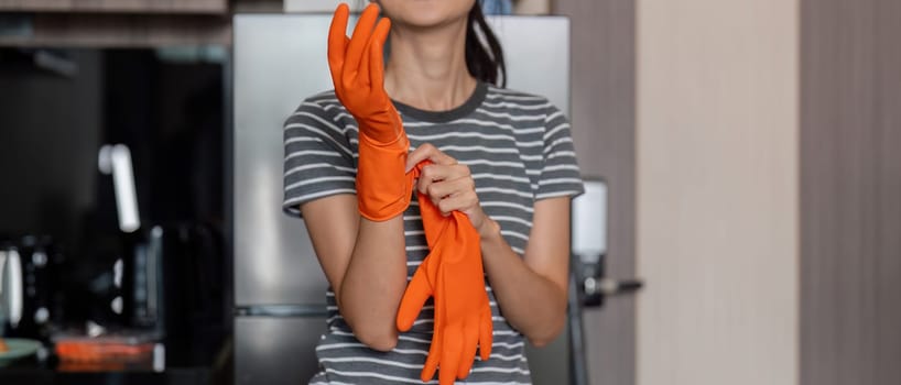 Dedicated young woman gearing up for house cleaning, putting on vibrant orange gloves while standing in her stylish kitchen.