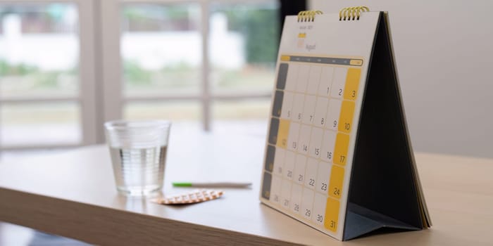 A monthly calendar with highlighted days, a glass of water, a pen, and a pack of contraceptive pills placed on a wooden table, emphasizing the importance of family planning.