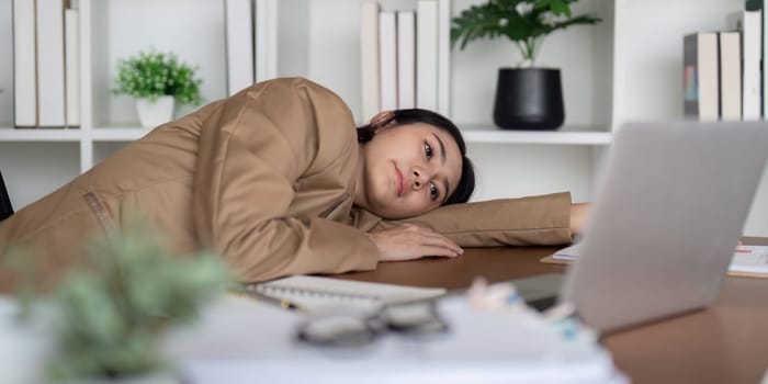 A businesswoman appears stressed and frustrated as she leans on her desk, surrounded by paperwork and her laptop, capturing a moment of challenge in her workday.