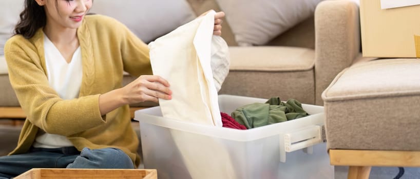 A happy woman folding items from a clear bin marked for donation, organizing clothes thoughtfully in her comfortable living room.