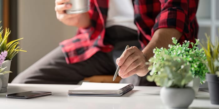 A young man in red checkered shirt focuses on writing notes in a notebook while holding coffee cup in stylish workspace with plants.