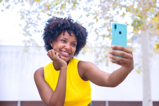 A joyful young woman with curly hair wearing a yellow top takes a selfie outdoors, smiling and gesturing peace. Vibrant scene showcasing happiness, individuality, and modern lifestyle.