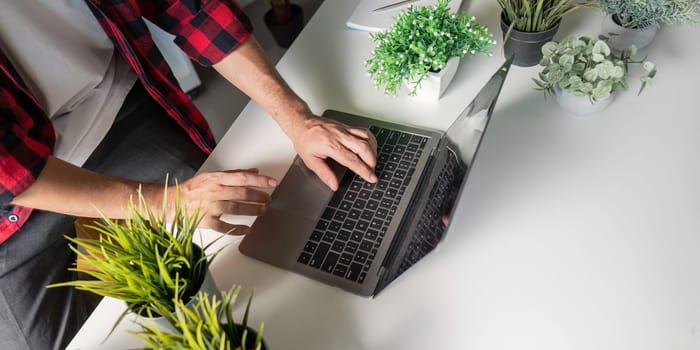 A close up view of a young man's hands on a laptop keyboard surrounded by lush greenery, illustrating the harmony between technology and plant care in an indoor setting.
