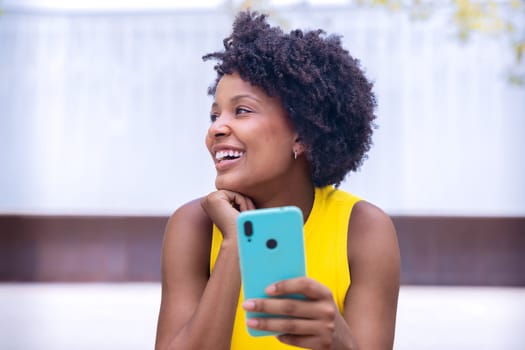 A young woman with natural curly hair wearing a yellow top rests her face on her hand while holding a smartphone. Calm and reflective moment captured in a bright outdoor setting.
