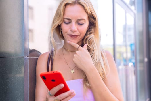 A blonde woman standing outdoors, focused on her smartphone. Captured in a modern urban environment, highlighting casual elegance, digital connectivity, and a relaxed lifestyle.