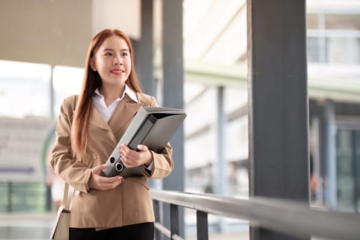 A poised businesswoman in a beige blazer smiles while walking through a contemporary office corridor, carrying important files with purpose.