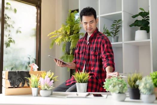 A young man in a flannel shirt thoughtfully examines his indoor plants using a tablet, showcasing his commitment to gardening in a bright and organized environment.