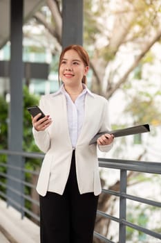 A happy businesswoman in a tailored white blazer stands confidently with a smartphone in one hand and files in the other, surrounded by a sleek office environment.