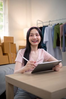 Happy young woman with braces confidently managing her online clothing store, holding a tablet in her inviting home workspace surrounded by packages.