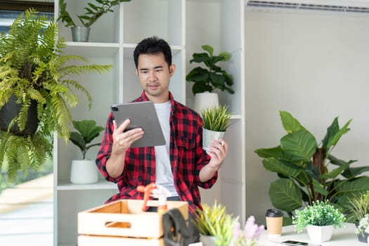 A young man in a flannel shirt examines a potted plant while looking at his tablet, highlighting his enthusiasm for indoor gardening and care in a bright setting.