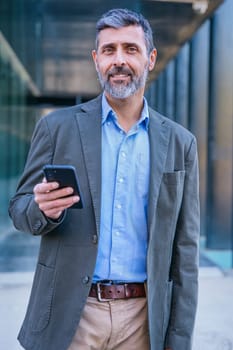 Smiling middle-aged businessman in a blue shirt holding and using a smartphone outdoors in a modern urban setting.