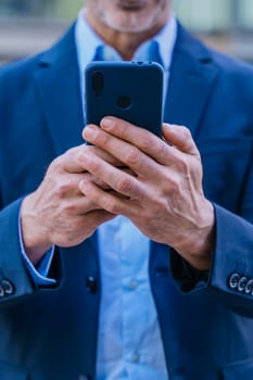 Smiling middle-aged businessman in a blue suit holding and using a smartphone outdoors in a modern urban setting.