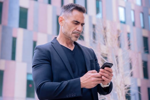 Smiling middle-aged businessman in a blue shirt holding and using a smartphone outdoors in a modern urban setting.