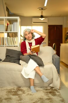 Happy an elderly woman sitting on a cozy sofa with a notebook and pen in hand.