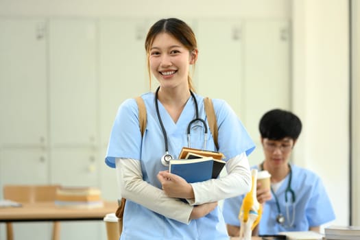 Smiling medical student in blue scrubs carrying textbooks standing in a classroom.