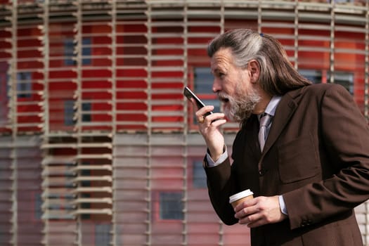 middle age male with beard in elegant suit with cup of takeaway coffee in hand talking on mobile phone against urban building, concept of business and mature man lifestyle, copyspace for text
