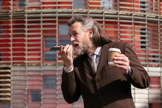 middle age male with beard in elegant suit with cup of takeaway coffee in hand recording voice message on mobile phone against urban building, concept of business and mature man lifestyle