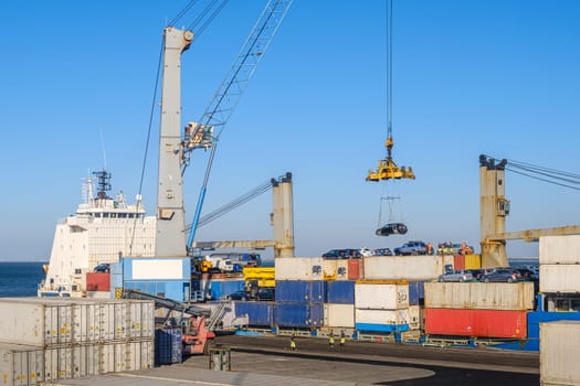 Large port crane loading car onto bulk carrier at shipping port. Colorful shipping containers stacked, ship-to-shore crane, shipment, reach stacker in operation, vehicles on top of freight containers, clear blue sky and sunny day, cargo terminal.
