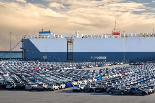 Large vehicle carrier ship known as RO-RO at port on sunny day, large parking area filled with cars waiting loading and transportation to buyers and car dealers. Blue sky and colorful clouds, vehicle transportation.
