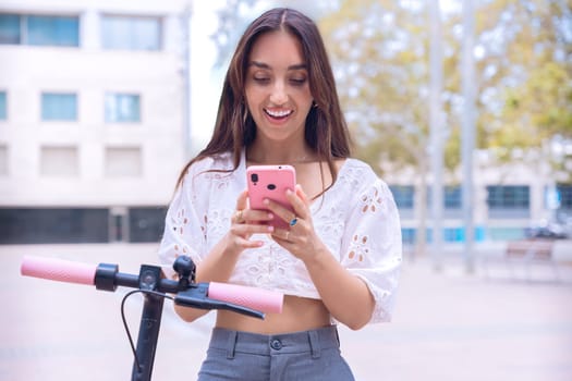 smiling young woman in a stylish white top uses her smartphone while standing next to an electric scooter in an urban setting. She appears excited and engaged, enjoying modern technology and mobility.