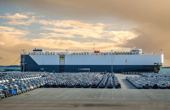 Large vehicle or car carrier ship known as RO-RO, roll on roll off, at port on sunny day, large parking area filled with cars. Blue sky and colorful clouds, vehicle transportation.