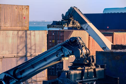 Reach stacker arms and spreaders maneuver while grabbing and carrying cargo containers in sea port shipping terminal on sunny day, clear blue sky.