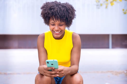 A young woman with natural curly hair wearing a yellow top rests her face on her hand while holding a smartphone. Calm and happy moment captured in a bright outdoor setting.