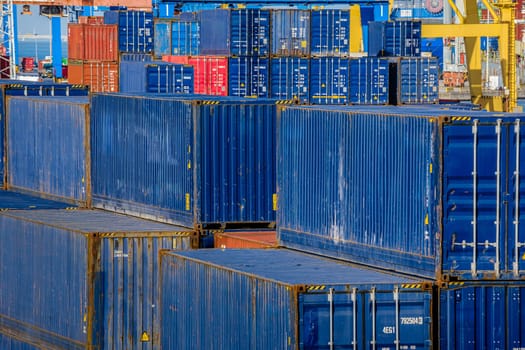 Close view of rows of blue cargo containers stacked at docking station in sunny day. Cranes in the background ready to load and unload cargo, maritime trade, shipping yard, storage, global logistics, freight.
