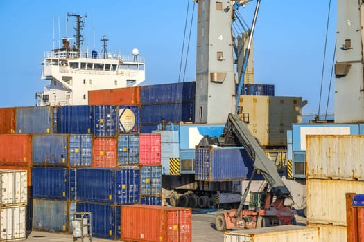 Reach stacker carry container for loading and unloading operations along bulk carrier in cargo port on sunny day. Clear blue skies, maritime logistics, cranes and cargo containers in shipping port, large vessel docked.