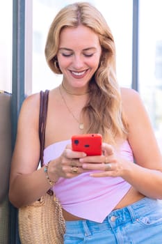 A blonde woman standing outdoors, focused on her smartphone. Captured in a modern urban environment, highlighting casual elegance, digital connectivity, and a relaxed lifestyle.